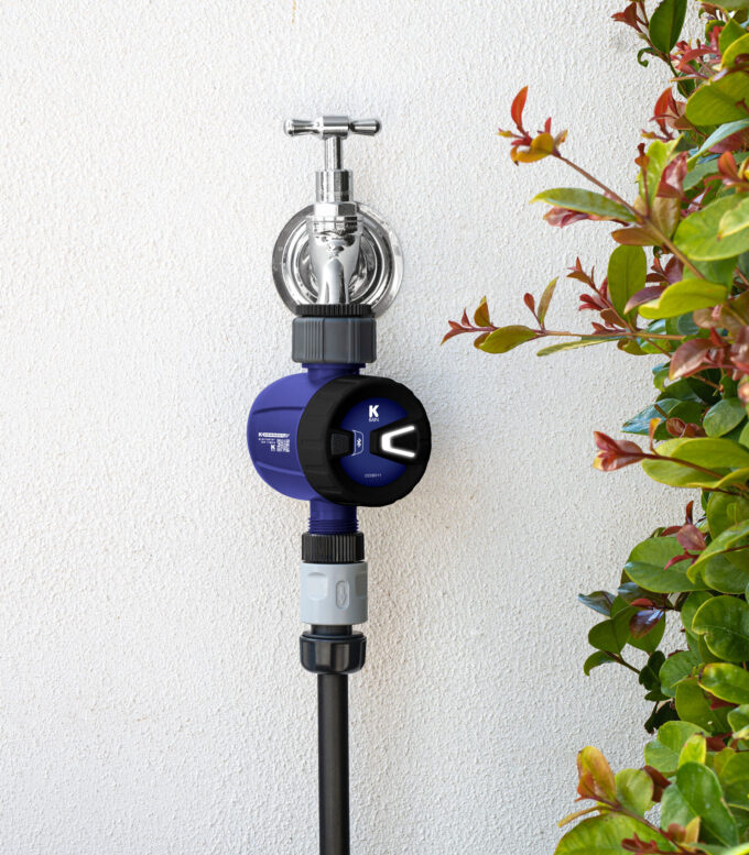 K Connect Bluetooth® Tap Timer connected to garden tap against a white stucco wall. A hedge of plants grows beside it.