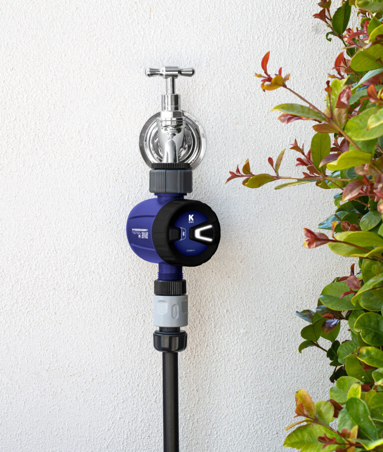 K Connect Bluetooth® Tap Timer connected to garden tap against a white stucco wall. A hedge of plants grows beside it.